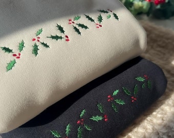 May include: A beige sweatshirt and a black sweatshirt stacked on top of each other. Both garments feature embroidered green holly leaves and red berries arranged in a horizontal line across the chest area, creating a festive holiday pattern.