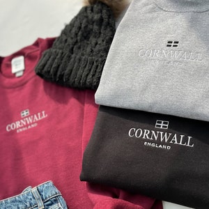 May include: Three sweatshirts with the text "Cornwall England" embroidered on the front. The sweatshirts are in burgundy, black, and gray. A black beanie and blue jeans are also in the image.