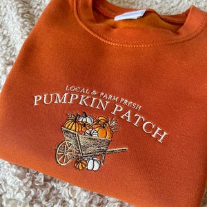 K&ouml;nnte beinhalten: Ein orangefarbenes Sweatshirt mit dem gestickten Text "LOCAL & FARM FRESH PUMPKIN PATCH". Das Design zeigt eine Holzkarre voller K&uuml;rbisse. Das Sweatshirt ist auf einer wei&szlig;en, strukturierten Oberfl&auml;che gefaltet.