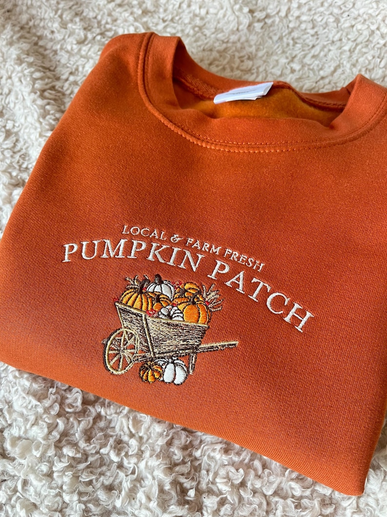K&ouml;nnte beinhalten: Ein orangefarbenes Sweatshirt mit dem gestickten Text "LOCAL & FARM FRESH PUMPKIN PATCH". Das Design zeigt eine Holzkarre voller K&uuml;rbisse. Das Sweatshirt ist auf einer wei&szlig;en, strukturierten Oberfl&auml;che gefaltet.
