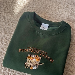 K&ouml;nnte beinhalten: Dunkelgr&uuml;nes Sweatshirt mit gesticktem Text und Grafik. Der Text lautet "LOCAL & FARM FRESH PUMPKIN PATCH" &uuml;ber einer Schubkarre voller K&uuml;rbisse und K&uuml;rbisse. Das Etikett lautet "Laura Schull".