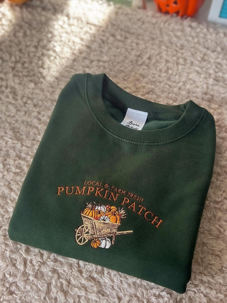 K&ouml;nnte beinhalten: Dunkelgr&uuml;nes Sweatshirt mit gesticktem Text und Grafik. Der Text lautet "LOCAL & FARM FRESH PUMPKIN PATCH" &uuml;ber einer Schubkarre voller K&uuml;rbisse und K&uuml;rbisse. Das Etikett lautet "Laura Schull".