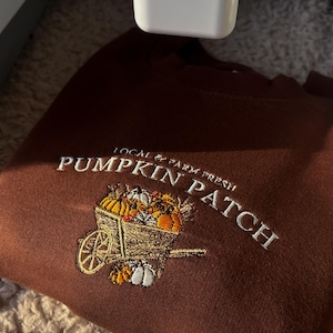 K&ouml;nnte beinhalten: Ein braunes Sweatshirt mit wei&szlig;er Stickerei, die "Pumpkin Patch" und "Local & Farm Fresh" zeigt. Das Design zeigt eine Schubkarre voller K&uuml;rbisse in verschiedenen Farben. Das Sweatshirt ist gefaltet.