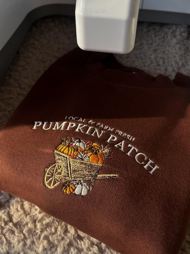 K&ouml;nnte beinhalten: Ein braunes Sweatshirt mit wei&szlig;er Stickerei, die "Pumpkin Patch" und "Local & Farm Fresh" zeigt. Das Design zeigt eine Schubkarre voller K&uuml;rbisse in verschiedenen Farben. Das Sweatshirt ist gefaltet.