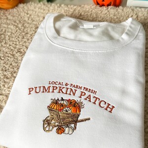 K&ouml;nnte beinhalten: Wei&szlig;es Sweatshirt mit orangefarbener Stickerei "PUMPKIN PATCH" und einer Schubkarre voller K&uuml;rbisse. Der Text "LOCAL & FARM FRESH" steht dar&uuml;ber. Das Sweatshirt ist gefaltet.