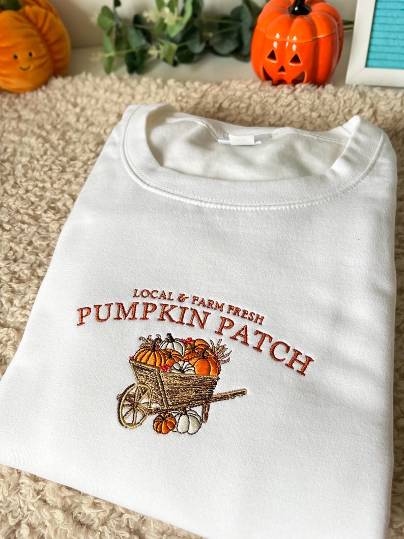 K&ouml;nnte beinhalten: Wei&szlig;es Sweatshirt mit orangefarbener Stickerei "PUMPKIN PATCH" und einer Schubkarre voller K&uuml;rbisse. Der Text "LOCAL & FARM FRESH" steht dar&uuml;ber. Das Sweatshirt ist gefaltet.