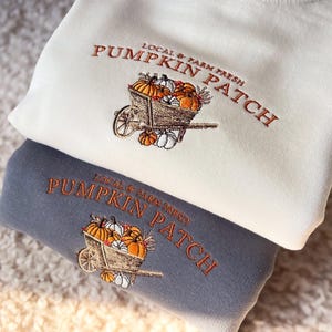 K&ouml;nnte beinhalten: Zwei Sweatshirts, eines cremefarbenes und eines graues, mit einem gestickten K&uuml;rbis-Patch-Design. Das Design zeigt eine Schubkarre voller K&uuml;rbisse und den Text "Local & Farm Fresh Pumpkin Patch" in Orange.