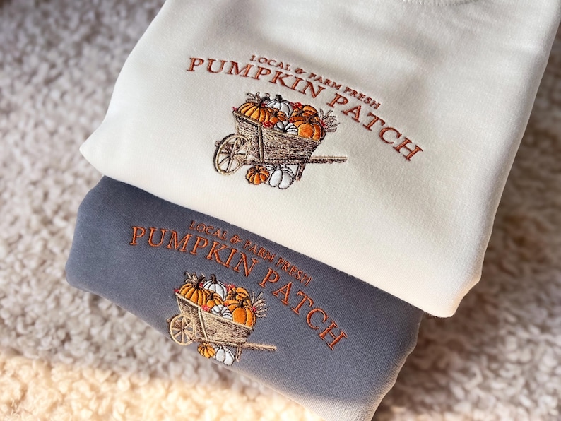 K&ouml;nnte beinhalten: Zwei Sweatshirts, eines cremefarbenes und eines graues, mit einem gestickten K&uuml;rbis-Patch-Design. Das Design zeigt eine Schubkarre voller K&uuml;rbisse und den Text "Local & Farm Fresh Pumpkin Patch" in Orange.