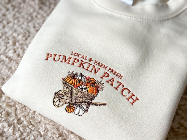 Embroidered sweatshirt with a Pumpkin Patch design, featuring pumpkins in a wagon with some autumn leaves on a cream sweater.