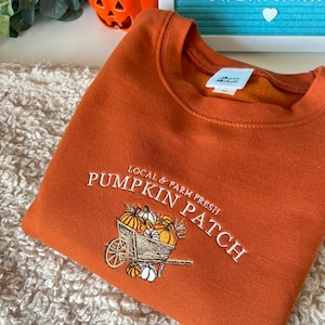 K&ouml;nnte beinhalten: Orangefarbenes Sweatshirt mit gesticktem "Pumpkin Patch"-Design, das einen Wagen voller K&uuml;rbisse zeigt. Der Text "Local & Farm Fresh" befindet sich dar&uuml;ber. Ein gem&uuml;tliches Kleidungsst&uuml;ck zum Thema Herbst.
