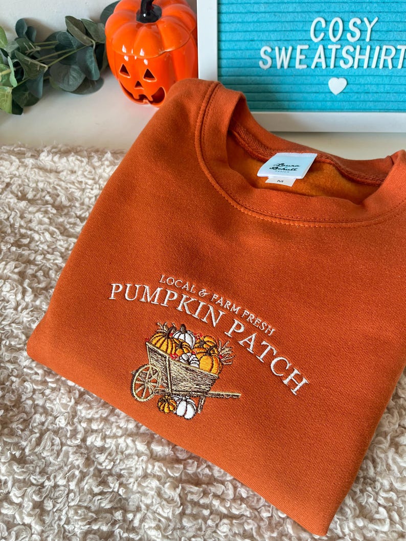 K&ouml;nnte beinhalten: Orangefarbenes Sweatshirt mit gesticktem "Pumpkin Patch"-Design, das einen Wagen voller K&uuml;rbisse zeigt. Der Text "Local & Farm Fresh" befindet sich dar&uuml;ber. Ein gem&uuml;tliches Kleidungsst&uuml;ck zum Thema Herbst.