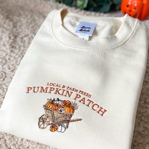 Embroidered sweatshirt with a Pumpkin Patch design, featuring pumpkins in a wagon with some autumn leaves on a cream sweater.