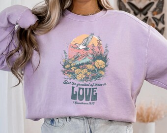 Puede incluir: Una sudadera de color lavanda con un estampado gráfico de un pájaro volando sobre una cordillera con flores. El texto "But the greatest of these is LOVE 1 Corinthians 13:13" está impreso debajo del gráfico.