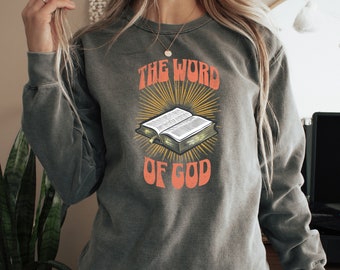 Puede incluir: Una sudadera gris con un gráfico de estilo vintage de un libro abierto con el texto "THE WORD OF GOD" en naranja.