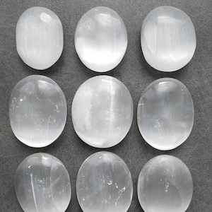 May include: Nine smooth, white selenite stones, each with a slightly oval shape, are arranged in three rows on a gray surface.