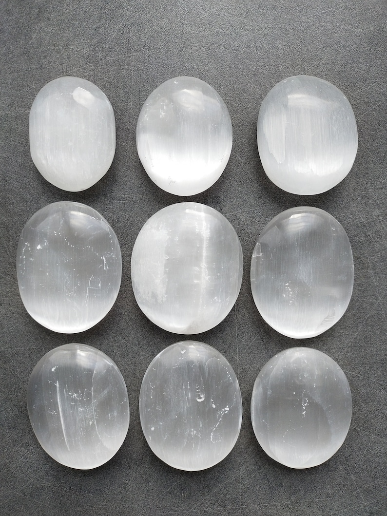 May include: Nine smooth, white selenite stones, each with a slightly oval shape, are arranged in three rows on a gray surface.