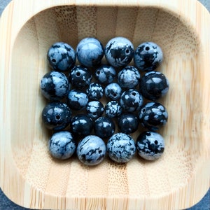 Op de afbeelding: Een houten kom gevuld met ronde kralen met zwarte en witte vlekken. De kralen zijn glad en hebben een natuurlijke, organische uitstraling.