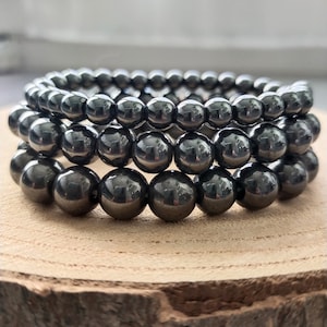 Puede incluir: Tres pulseras apiladas hechas de cuentas de hematita negra. Las pulseras están sobre una superficie de madera.