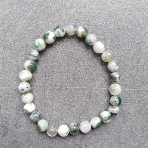 Könnte beinhalten: Ein Armband mit runden Moosachat-Perlen in Grün-, Weiß- und transparenten Farbtönen. Die Perlen sind auf einem elastischen Band aufgereiht. Jede Perle hat einen Durchmesser von etwa 0,8 cm. Das Armband liegt auf einer grauen Oberfläche.