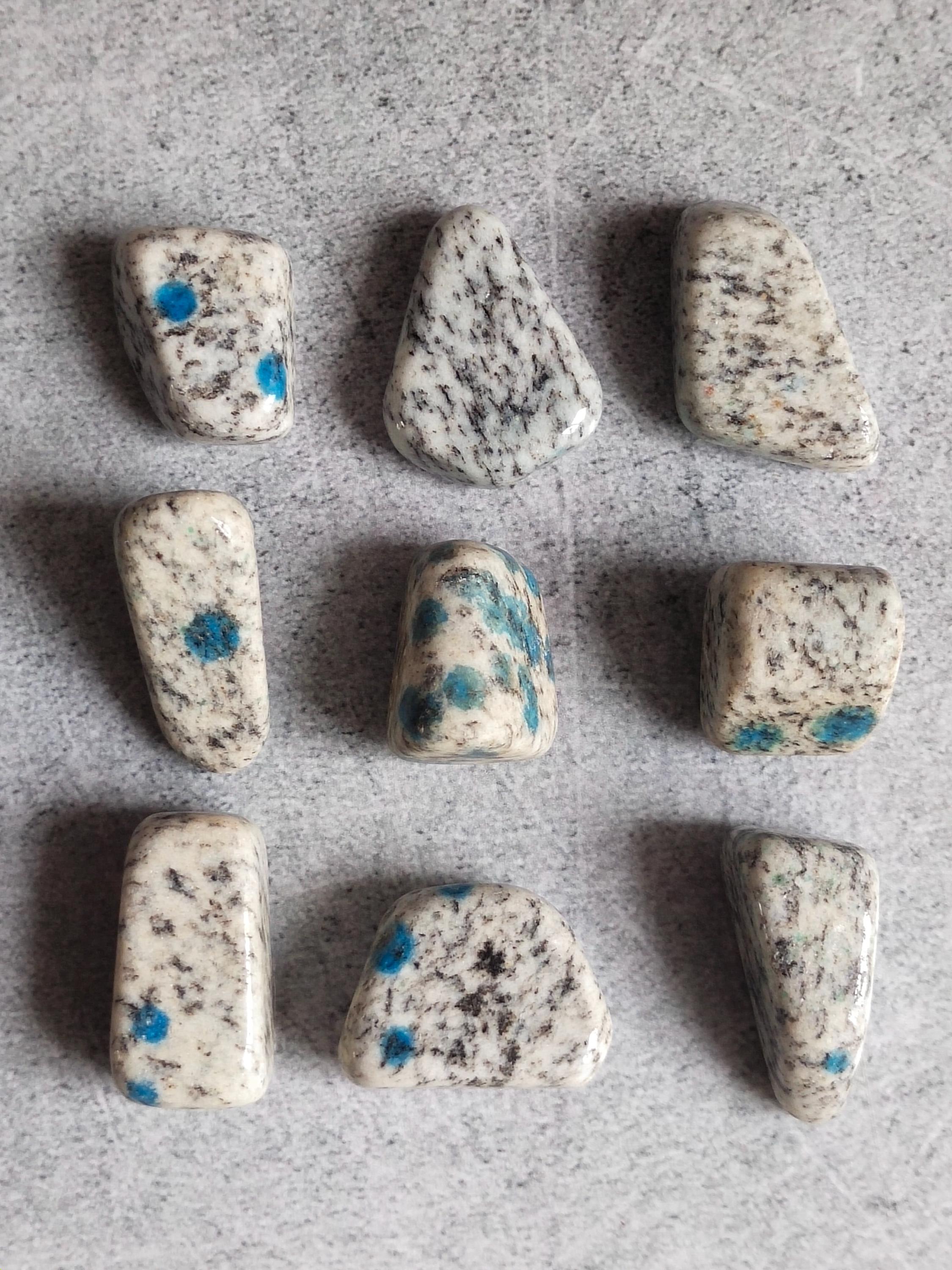 May include: Nine polished K2 stones, also known as K2 jasper, are displayed. The stones have a white base with black speckling and vibrant blue spots. The shapes vary, including rectangular, triangular, and rounded forms. The stones are arranged on a light gray surface.