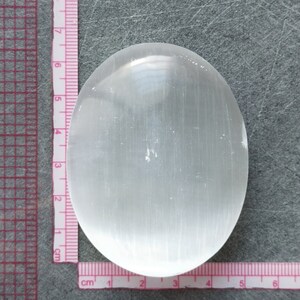 May include: A smooth, white selenite palm stone with a slightly oval shape. The stone is approximately 6 cm long and 4 cm wide.