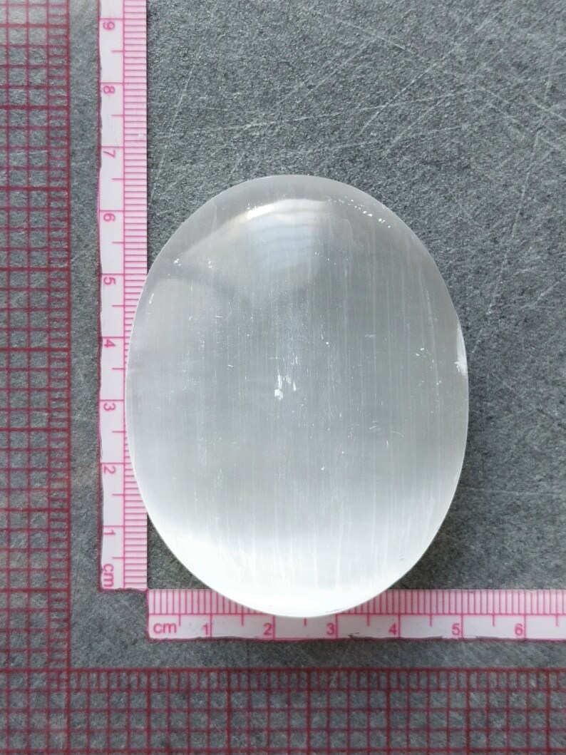 May include: A smooth, white selenite palm stone with a slightly oval shape. The stone is approximately 6 cm long and 4 cm wide.
