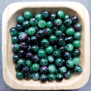 Peut inclure: Un bol rempli de perles de pierres précieuses vertes, noires et roses. Les perles sont rondes et ont une finition lisse et polie.