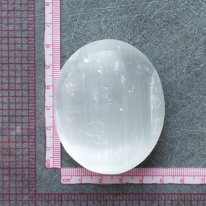 May include: A white selenite palm stone with a smooth, polished surface and visible striations. The stone is approximately 5 cm in diameter.