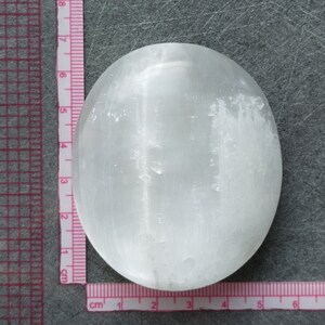 May include: A white selenite palm stone, measuring approximately 6 cm in diameter, with a smooth, polished surface and visible striations.