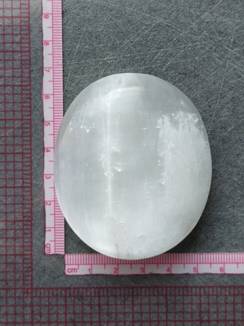 May include: A white selenite palm stone, measuring approximately 6 cm in diameter, with a smooth, polished surface and visible striations.