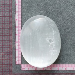 May include: A smooth, white selenite palm stone with a slightly oval shape. The stone is approximately 6 cm long and 4 cm wide.