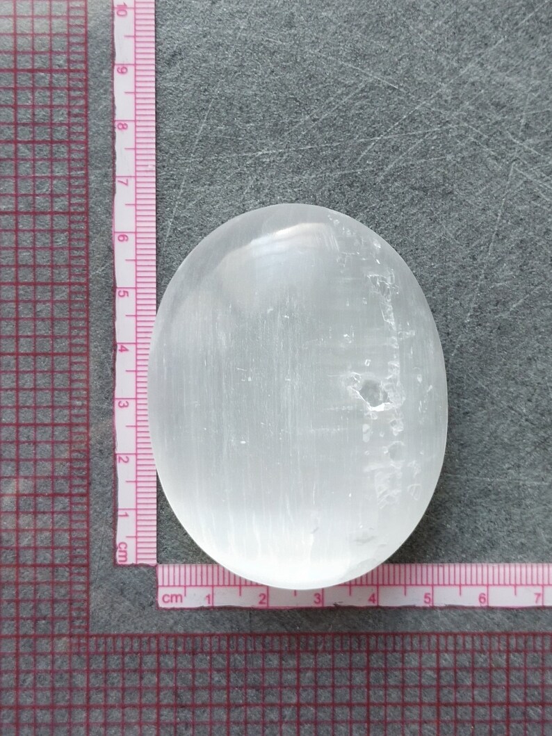 May include: A smooth, white selenite palm stone with a slightly oval shape. The stone is approximately 6 cm long and 4 cm wide.