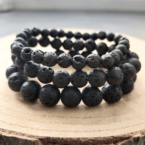 Peut inclure: Trois bracelets de perles en pierre de lave noire. Les bracelets sont faits de perles rondes et poreuses et sont empilés sur une surface en bois. Les bracelets sont de couleur charbon foncé et semblent faits à la main.