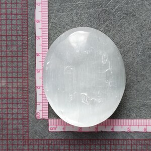 May include: A white selenite palm stone, approximately 5 cm in diameter, with a smooth, polished surface. The stone has a natural, cloudy appearance with subtle striations.