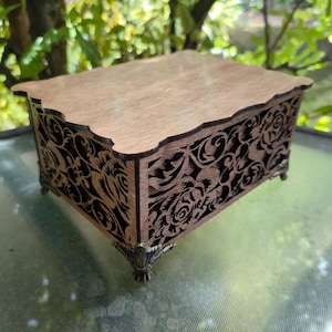 Op de afbeelding: Een houten sieradendoosje met ingewikkelde bloemmotieven. De doos heeft een lichtbruin deksel en donkerbruine zijkanten met gedetailleerde gravures. Het staat op kleine, decoratieve voetjes, met een mix van natuurlijke houttinten en sierlijk design.