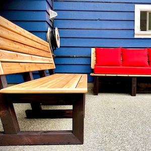 Könnte beinhalten: Eine Holzbank mit naturbelassenem Holzfinish und dunkelbraunen Beinen. Die Bank besteht aus Latten und hat ein schlichtes Design. Sie steht auf einer Betonterrasse vor einem blauen Haus.