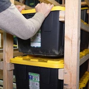 May include: A person placing a black and yellow storage bin on a wooden shelf. The bin has a yellow lid and a label that reads "Greenmade Storage 27 Gal. Professional Grade Box".