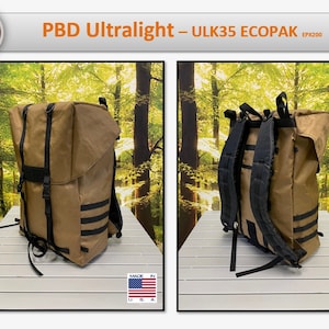 May include: A brown canvas backpack with black straps and accents. The backpack has a roll-top closure and is made in the USA. The backpack is shown in front of a blurred background of trees.