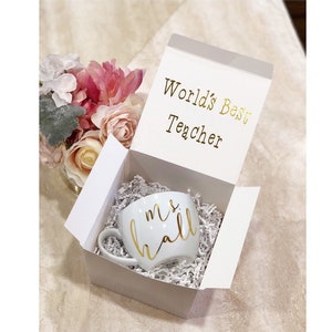 K&ouml;nnte beinhalten: Eine wei&szlig;e Tasse mit goldener Aufschrift "mrs hall" in einer wei&szlig;en Geschenkbox mit der Aufschrift "World's Best Teacher" auf dem Deckel.