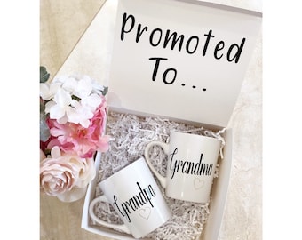 May include: White gift box with the text "Promoted To..." on the lid. Inside the box are two white mugs with black text. One mug says "Grandpa" with a small heart and the other says "Grandma" with a small heart.