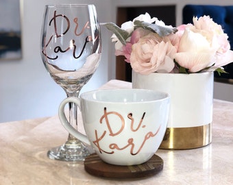 May include: A white ceramic mug with a gold rim and a rose gold design that says "Dr. Kara" on it. The mug is sitting on a wooden coaster. A wine glass with a rose gold design that says "Dr. Kara" is in the background.