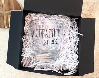 May include: A black gift box with white shredded paper inside. The box contains a clear glass with the text "GODFATHER EST. 2021" printed on it.