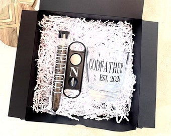 May include: A black gift box with white tissue paper inside. The box contains a glass with the text "Godfather Est. 2021", a cigar cutter, and a cigar tube.
