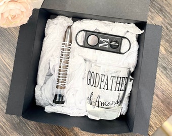 May include: A black gift box with a white tissue paper lining. The box contains a glass with the text "Godfather of Amanda", a cigar cutter, and a cigar tube.