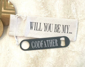 Pode incluir: Uma bolsa de tecido branca com o texto "Will you be my...Godfather" impresso nela. Um abridor de garrafas preto com a palavra "Godfather" está em cima da bolsa.