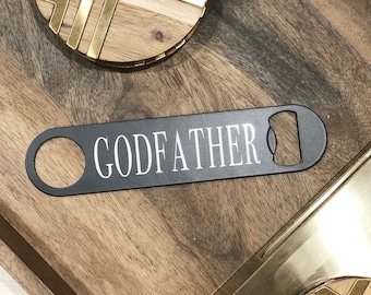 May include: Black metal bottle opener with the word "GODFATHER" printed in white. The bottle opener is on a wooden surface with a gold and wood coaster.