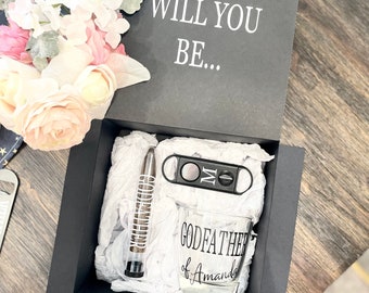 May include: A black gift box with a white card that says "Will You Be..."  The box contains a glass with the text "Godfather of Amanda", a cigar cutter, and a cigar.