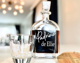 May include: A clear glass decanter with a black label that reads "Padrino de Ellie" and a matching clear glass tumbler. The decanter is filled with a dark liquid. A wooden coaster with a gold geometric design is in the background.