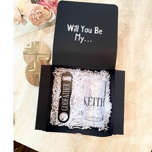 以下が含まれることがあります： 黒いギフトボックスに「Will You Be My... 」という白い文字が書かれています。ボックスが開いており、中には「Keith」と書かれたグラス、ボトルオープナー（「Godfather」の文字入り）、白いシュレッダーペーパーが入っています。