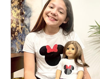 May include: A girl and a doll are wearing matching white t-shirts with a black and red Mickey Mouse silhouette. The doll is wearing colorful leggings.